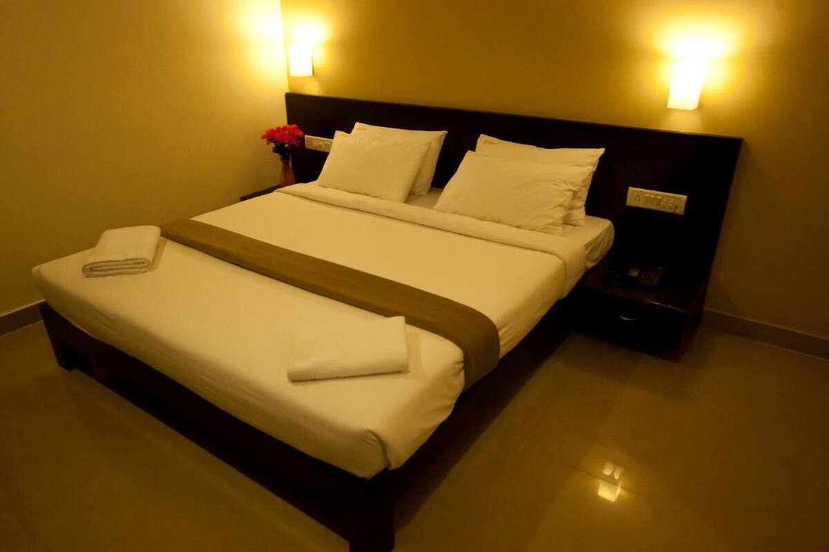 Una cama bien hecha con sábanas blancas y una colcha marrón, dos mesitas de noche con lámparas, y un florero con flores en el lado izquierdo.