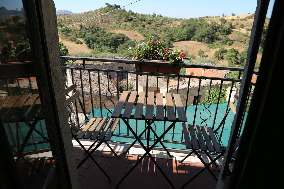 A balcony with a view of a hilly landscape, a wooden table, and a potted plant with flowers.