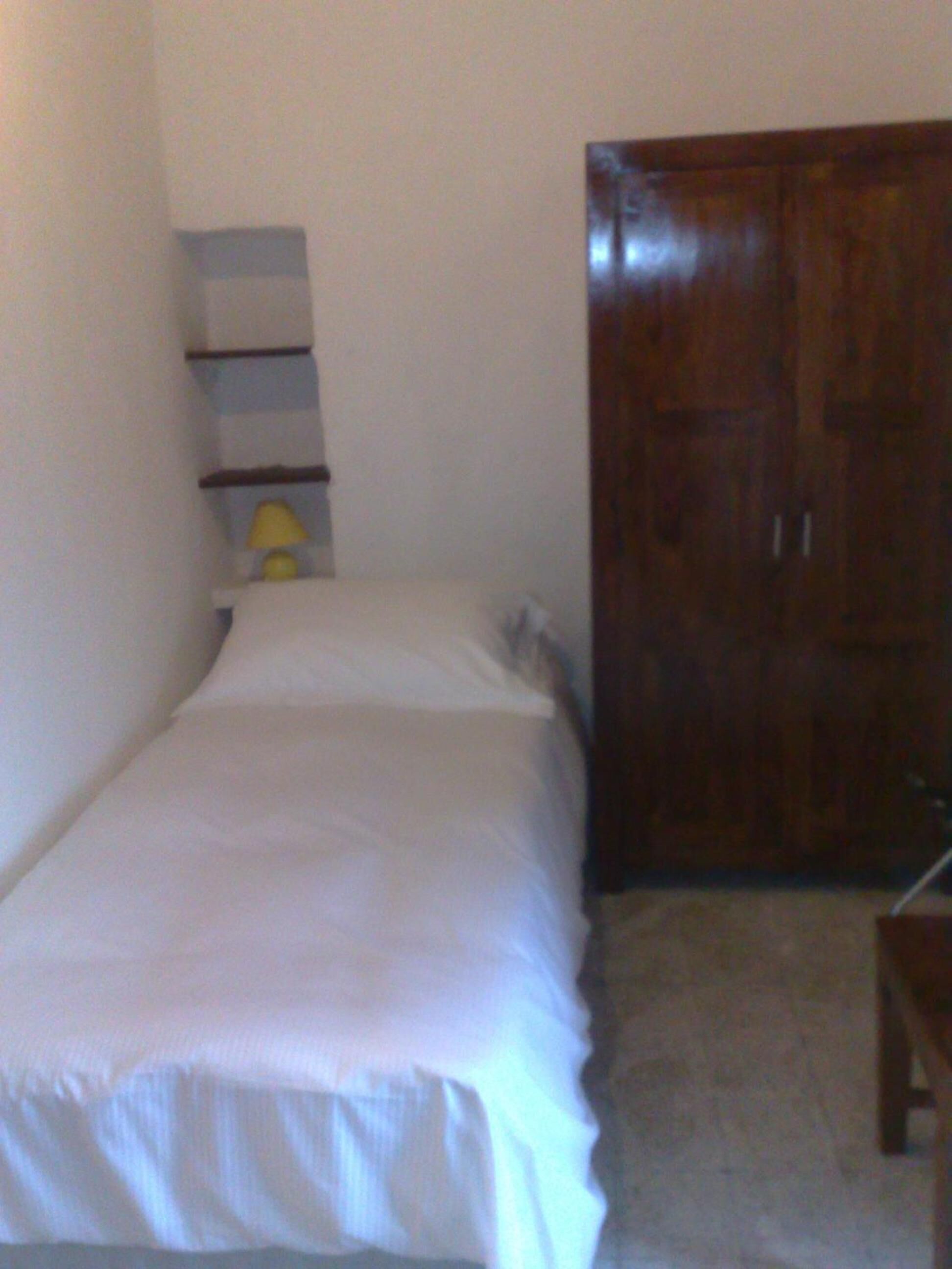 Une petite chambre avec un lit simple, une armoire en bois et une étagère fixée au mur.