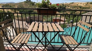 A balcony with a wooden table and chairs, a hanging flower pot, and a view of a swimming pool. - Locanda di Cadì (Petralia Soprana)