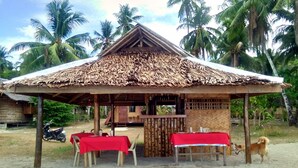 A rustic hut with a thatched roof, surrounded by palm trees. There are two tables with red tablecloths and wooden chairs. A dog is visible near the hut. - La Acuario Beach Inn (San Vicente)