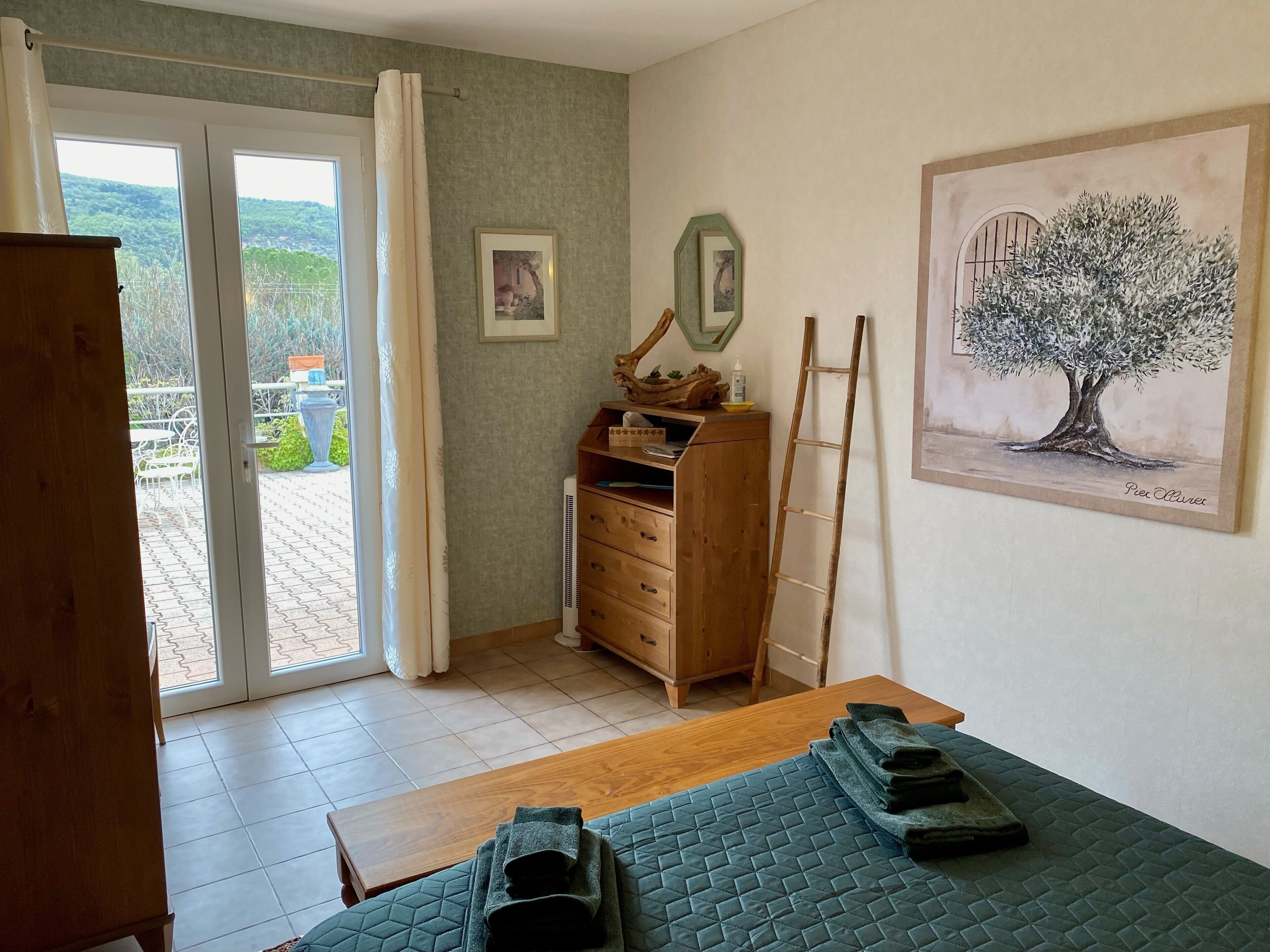Une pièce avec une commode en bois, un lit recouvert d’une courtepointe verte, une table en bois et une échelle appuyée contre le mur. Deux tableaux encadrés sont accrochés au mur, ainsi qu’une grande peinture représentant un arbre.