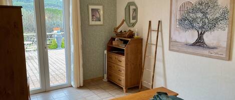 Une pièce avec une commode en bois, un lit recouvert d’une courtepointe verte, une table en bois et une échelle appuyée contre le mur. Deux tableaux encadrés sont accrochés au mur, ainsi qu’une grande peinture représentant un arbre.
