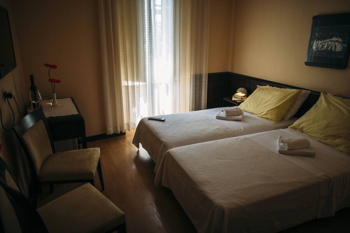 A hotel room with two beds, a desk with a lamp, a chair, and a window with curtains.