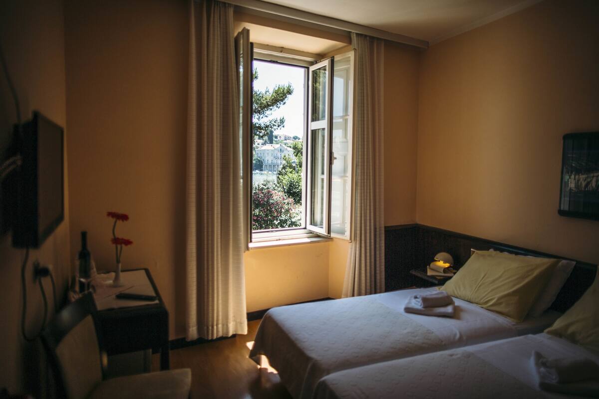 A hotel room with two beds, a window with curtains, a bedside table with a lamp, and a chair.
