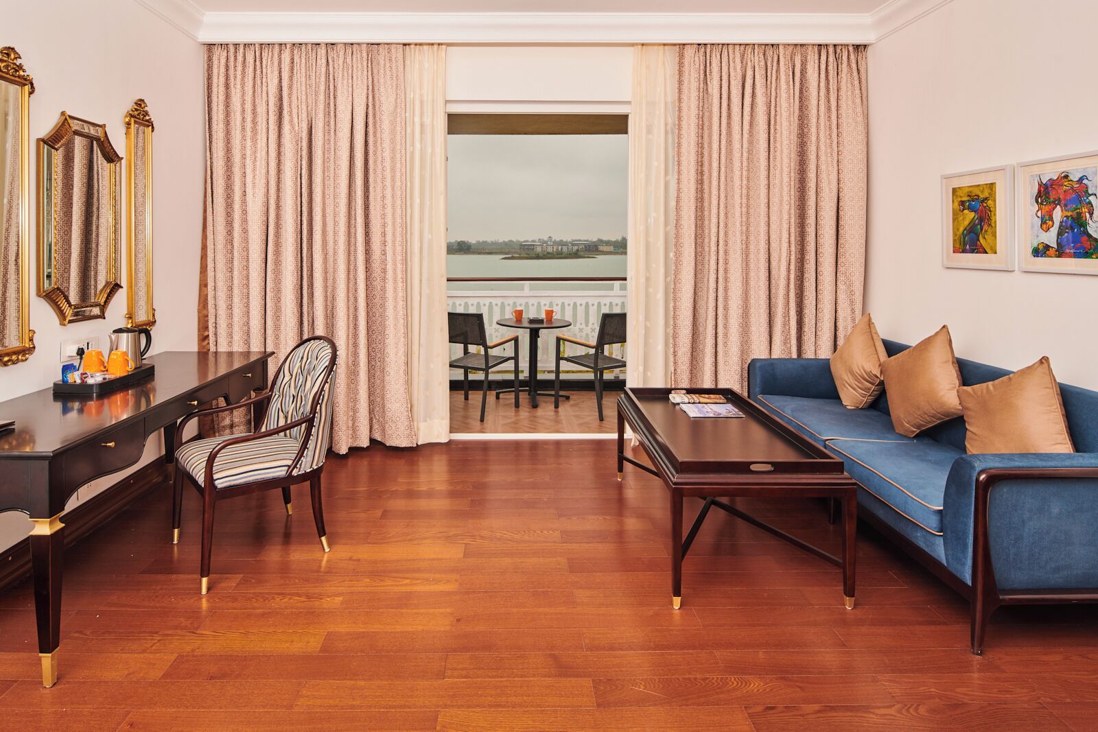 A hotel room with a wooden floor, a blue sofa, a wooden coffee table, a desk with a chair, and a view of a body of water through large windows.