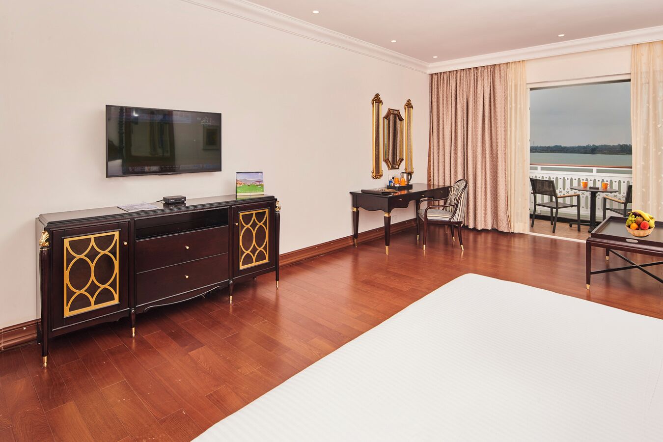 A hotel room with a large bed, a TV mounted on the wall, a desk with a chair, and a balcony with a view.