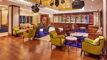 A spacious lobby with wooden flooring, multiple seating areas including armchairs and sofas, a bar with shelves of bottles, and a large window offering a view of the city.
