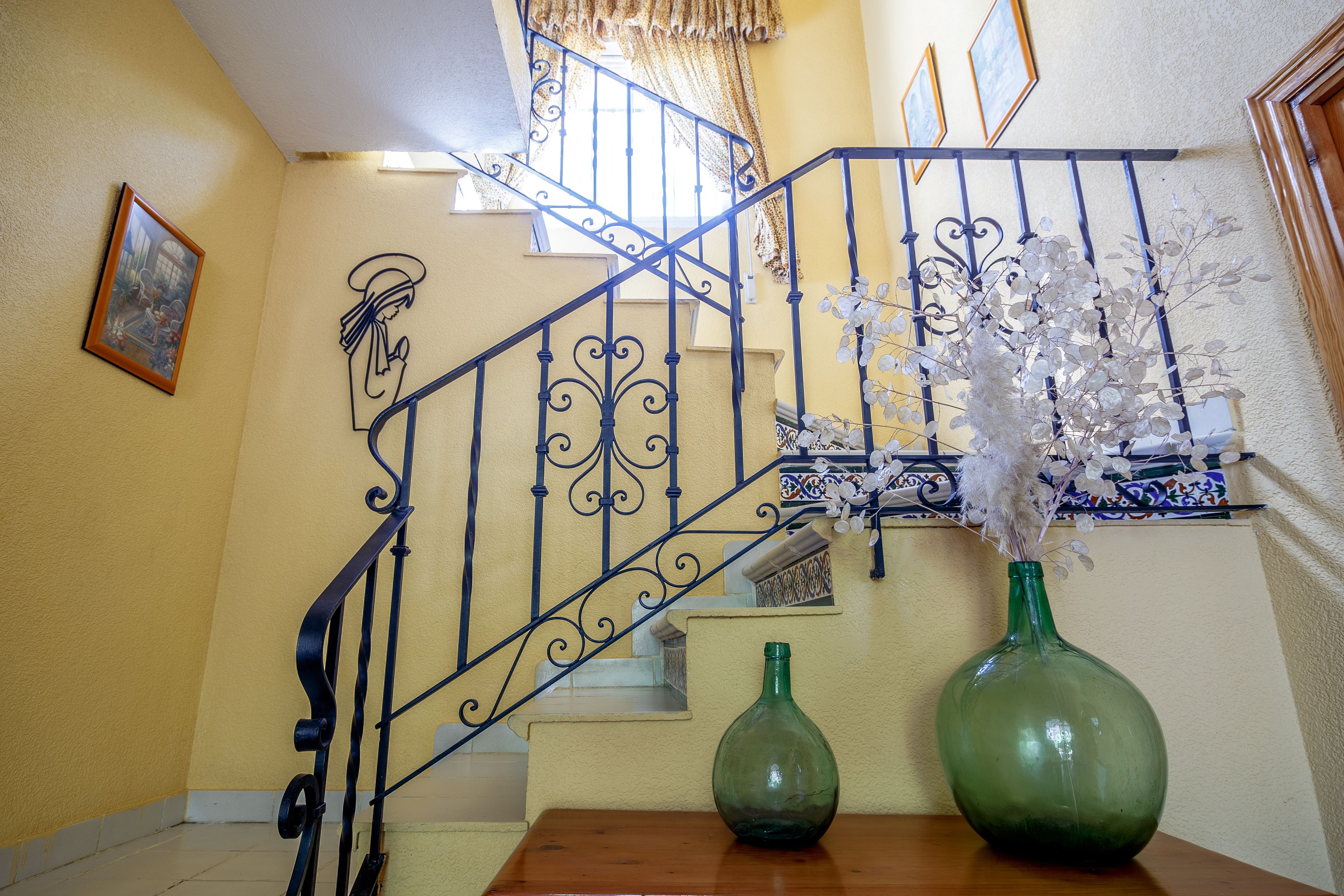 Una escalera con barandillas negras ornamentales, un jarrón verde con flores blancas sobre una mesa de madera y un cuadro en la pared.