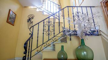 Una escalera con barandillas negras ornamentales, un jarrón verde con flores blancas sobre una mesa de madera y un cuadro en la pared.