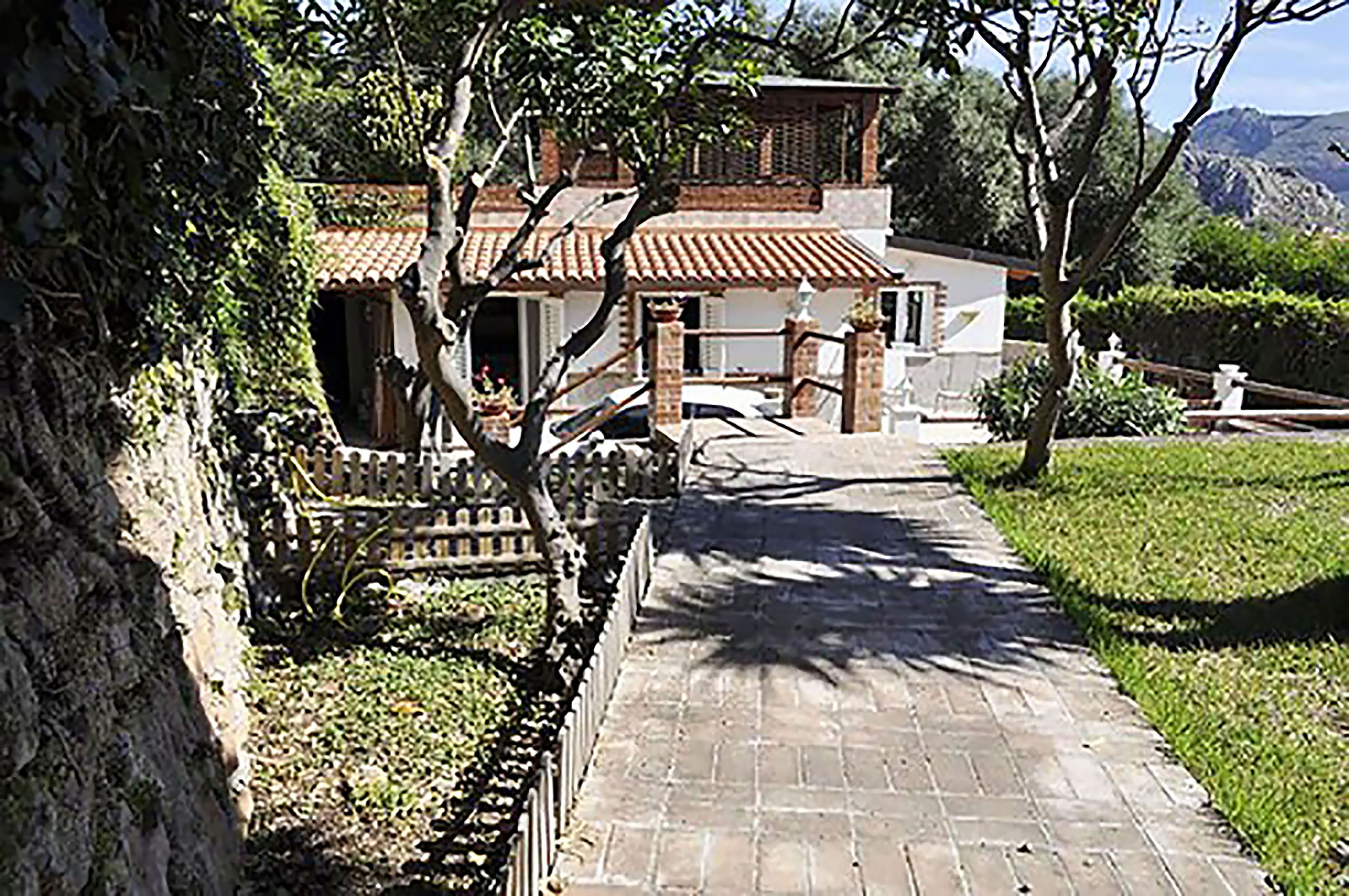Un sendero conduce a un edificio tradicional con techo de tejas, rodeado de follaje y un muro de piedra.