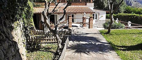 Un sendero conduce a un edificio tradicional con techo de tejas, rodeado de follaje y un muro de piedra.