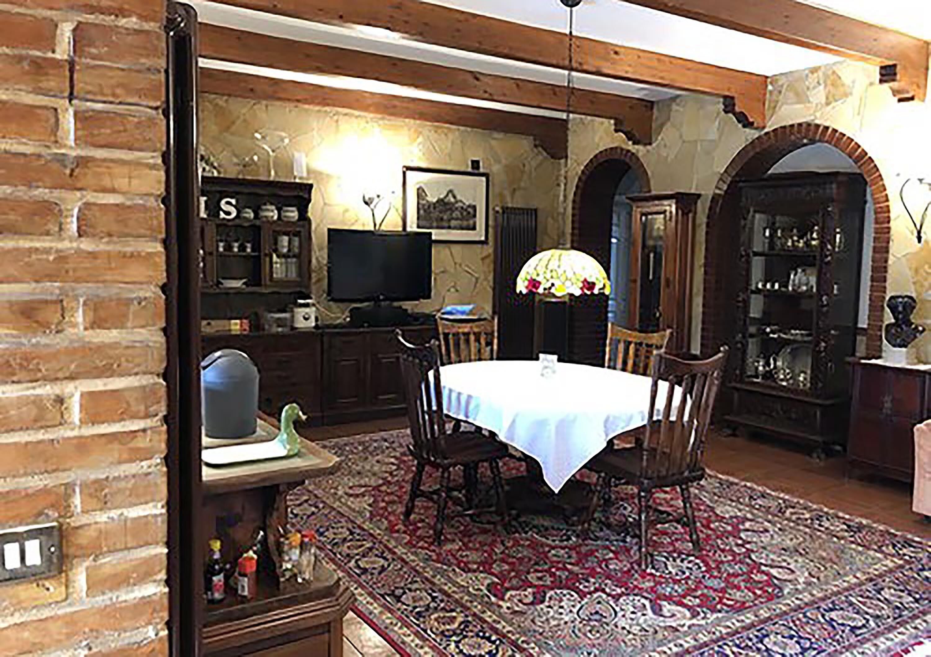 Un'area pranzo con un tavolo apparecchiato per quattro, circondato da sedie di legno. Si vede una televisione su un mobile, un lampadario e un tappeto decorato a pavimento.