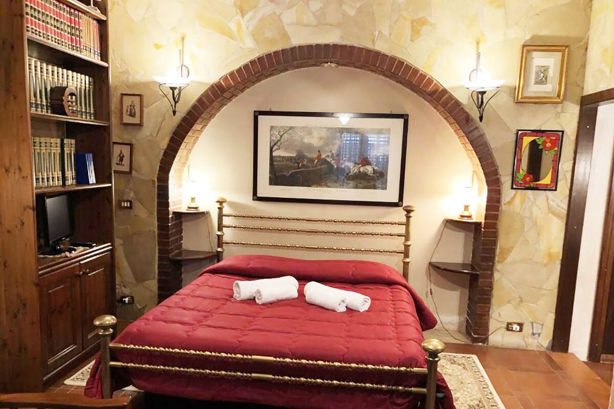 A hotel room with a red bed, a wooden bookshelf, and a painting on the wall.
