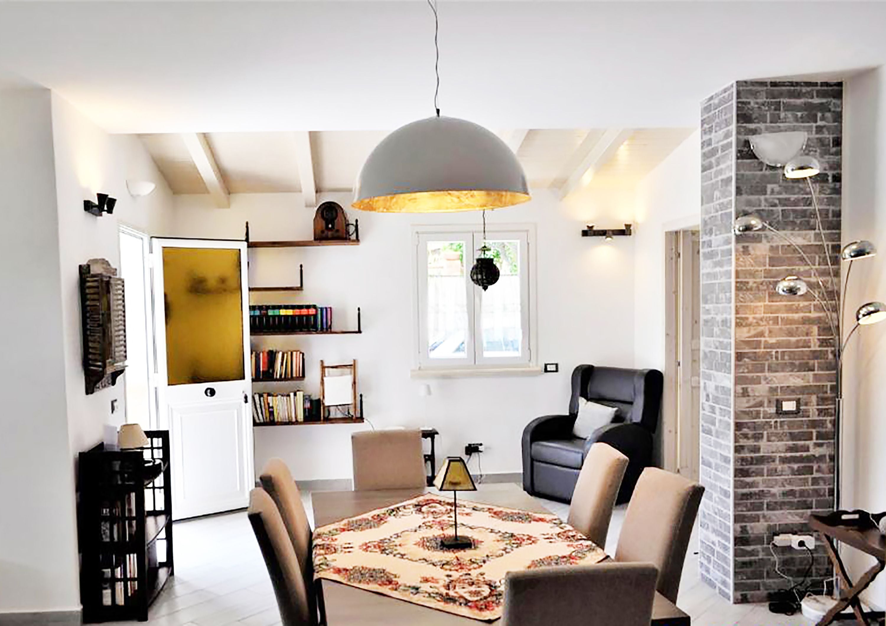 Een woonkamer met een ronde tafel, stoelen, een boekenkast, een open haard en een groot raam.