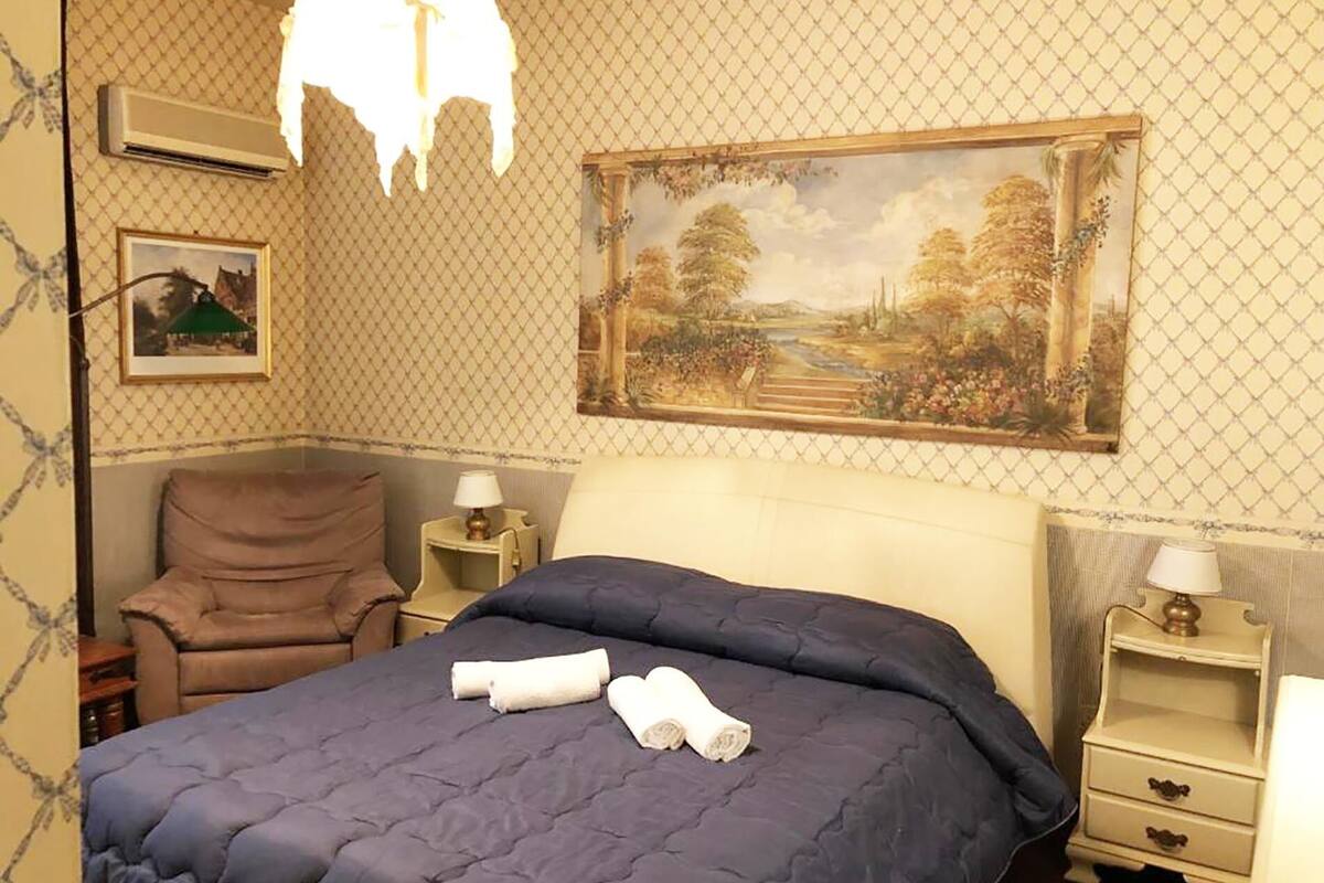 A hotel room with a large bed, a chair, a nightstand, a lamp, and a painting on the wall.