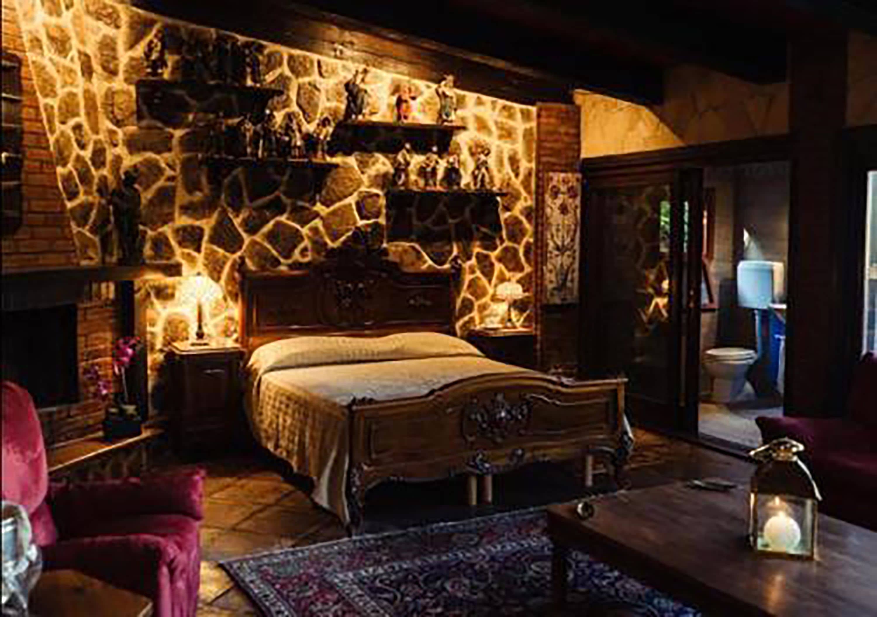 A bedroom with a stone wall, a bed, a nightstand, a lamp, and a small table with a candle.