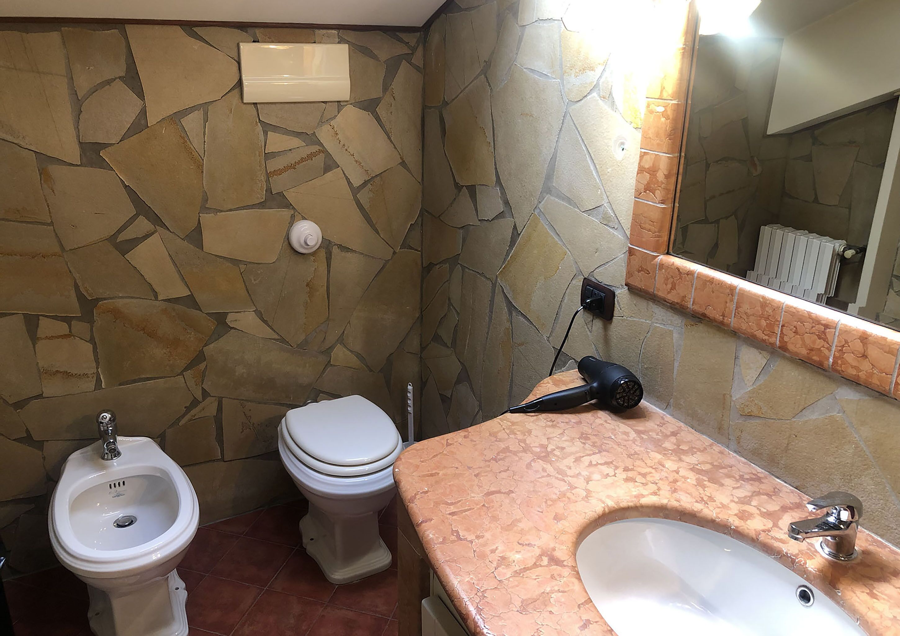 Un baño con pared de piedra, inodoro blanco, bidé y lavamanos con espejo.