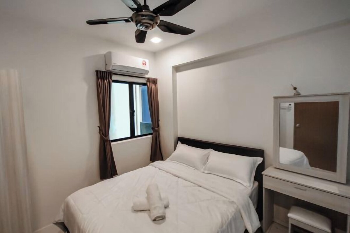 Habitación de hotel con cama, ventilador de techo, equipo de aire acondicionado, ventana con cortinas, espejo y una mesita.