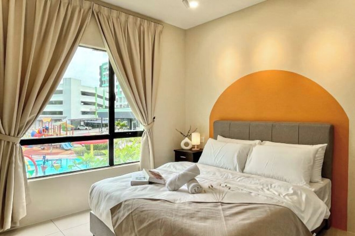 A hotel room with a bed, a nightstand, a lamp, and a view of a swimming pool and buildings.