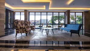 Un vestíbulo amplio con piso de mármol, ventanas grandes y mobiliario moderno, incluyendo un sofá azul y sillones estampados.