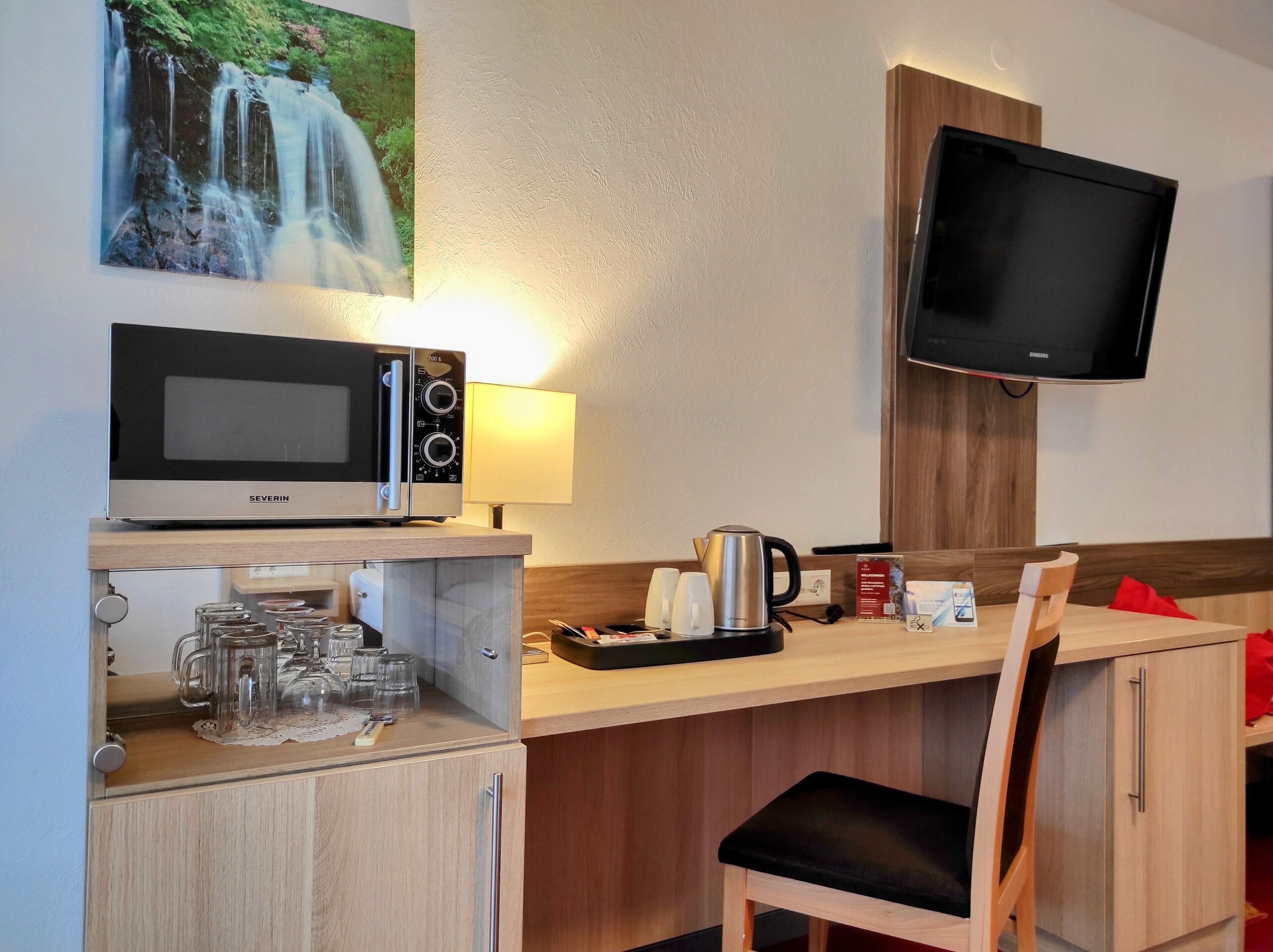 Una kitchenette equipada con microondas, hervidor de agua y cafetera, un televisor montado en la pared y una silla de madera.
