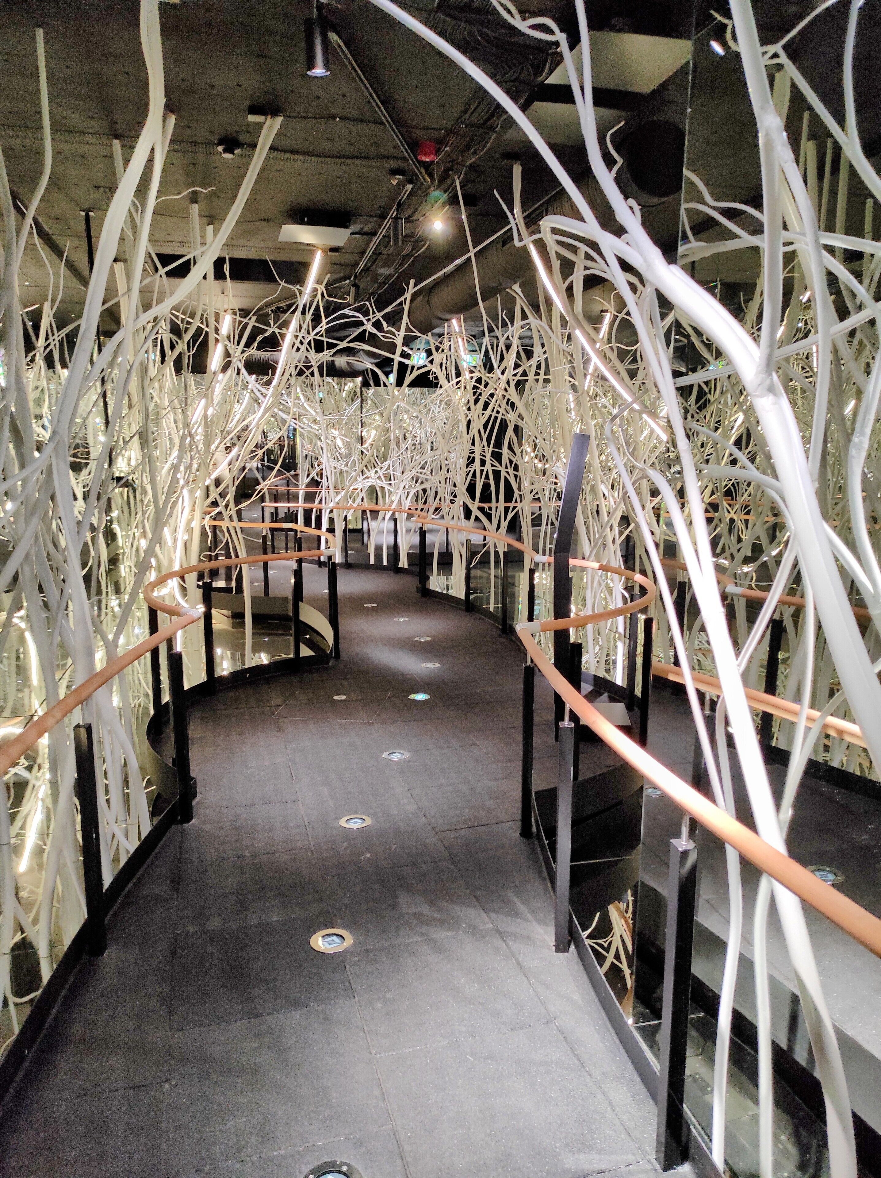 A corridor with a curved railing, white branches hanging from the ceiling, and a tiled floor.