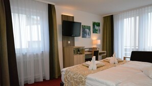 A hotel room with a large bed, a flat-screen TV mounted on the wall, a desk with a lamp, and a window with sheer curtains. - Wellness Hotel Garni Krone (Baiersbronn)