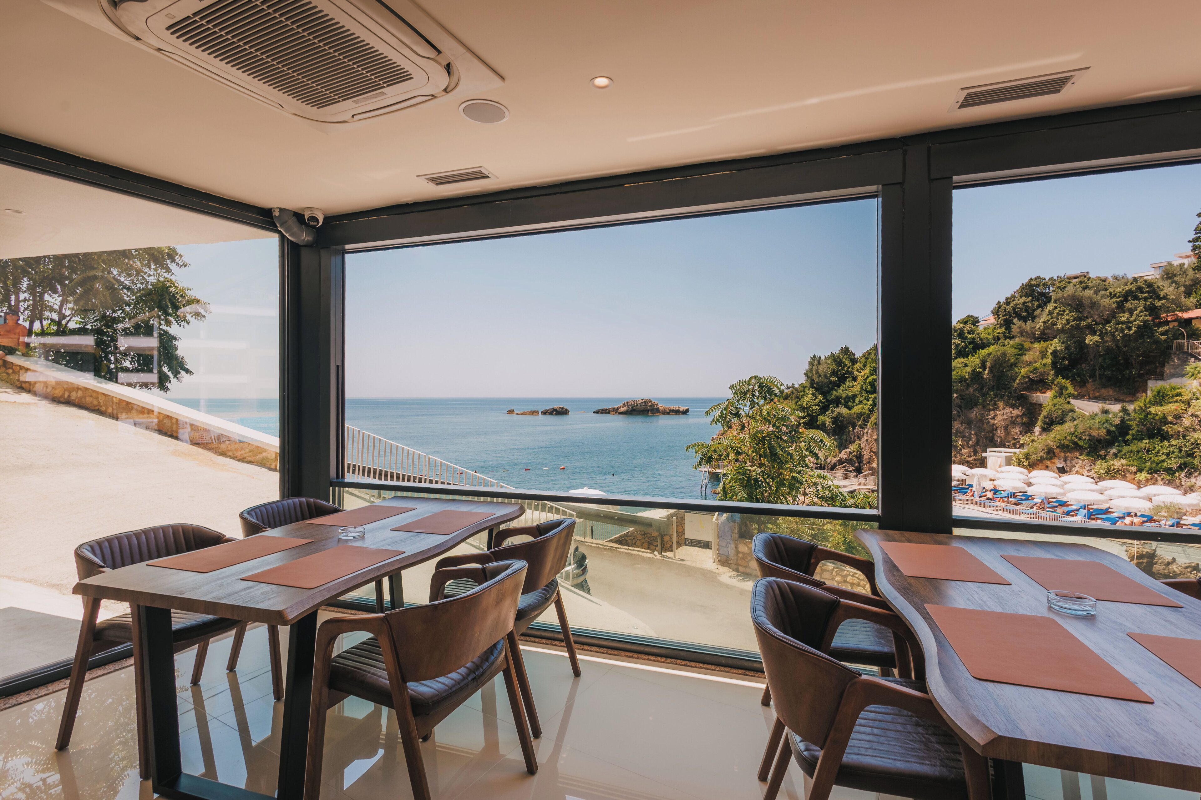 Un espace repas avec des tables et des chaises, de grandes fenêtres offrant une vue sur la mer et une côte rocheuse.
