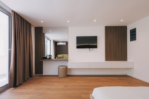 Quarto de hotel moderno com uma janela grande, televisão de tela plana na parede, um pequeno espaço de estar com um sofá e piso de madeira.