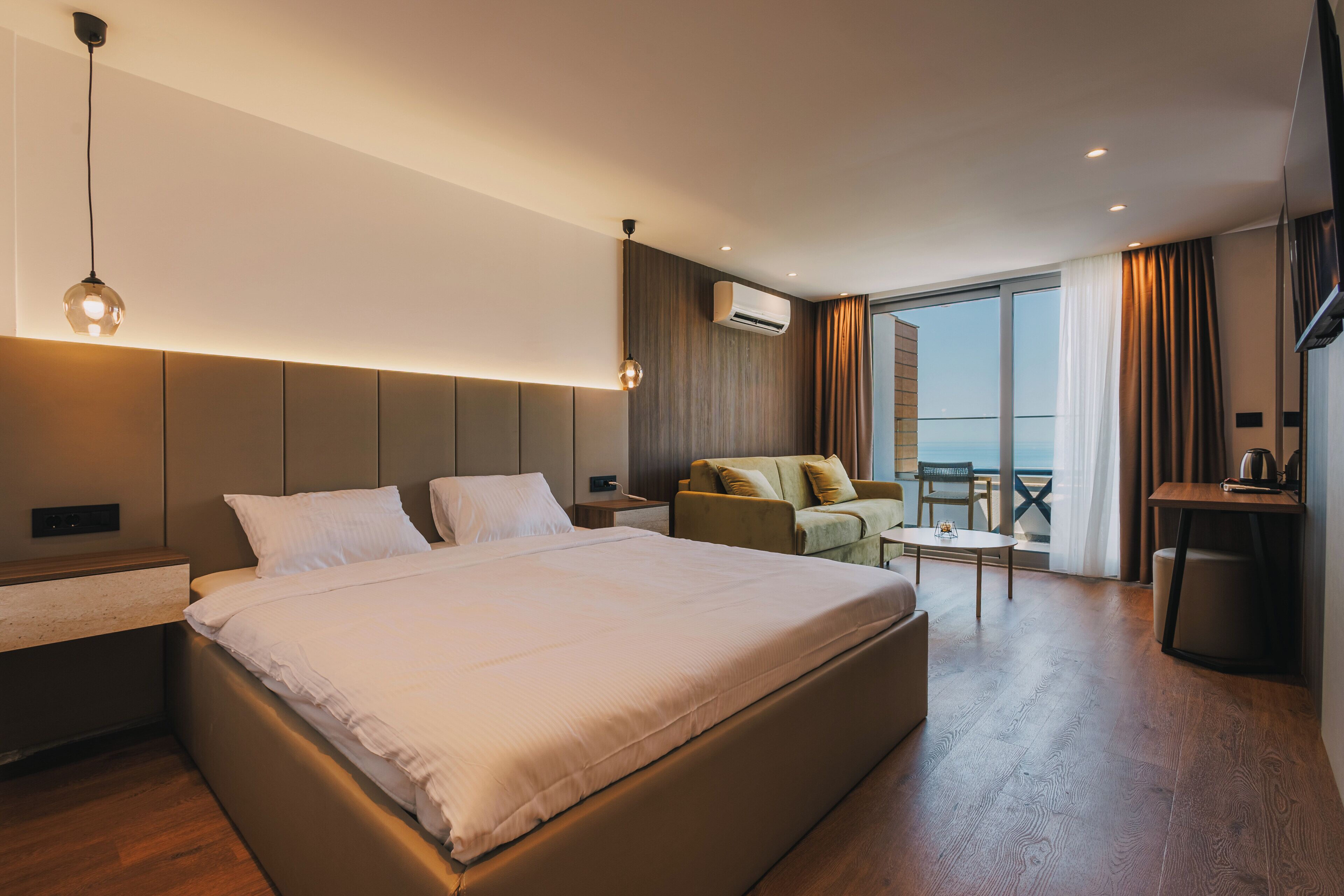 A modern hotel room with a large bed, a sofa, a small table, and a view of the sea.