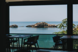 A view from inside a building with a large window overlooking a rocky coastline and calm sea. - Hotel Mediterraneo Liman (Ulcinj)