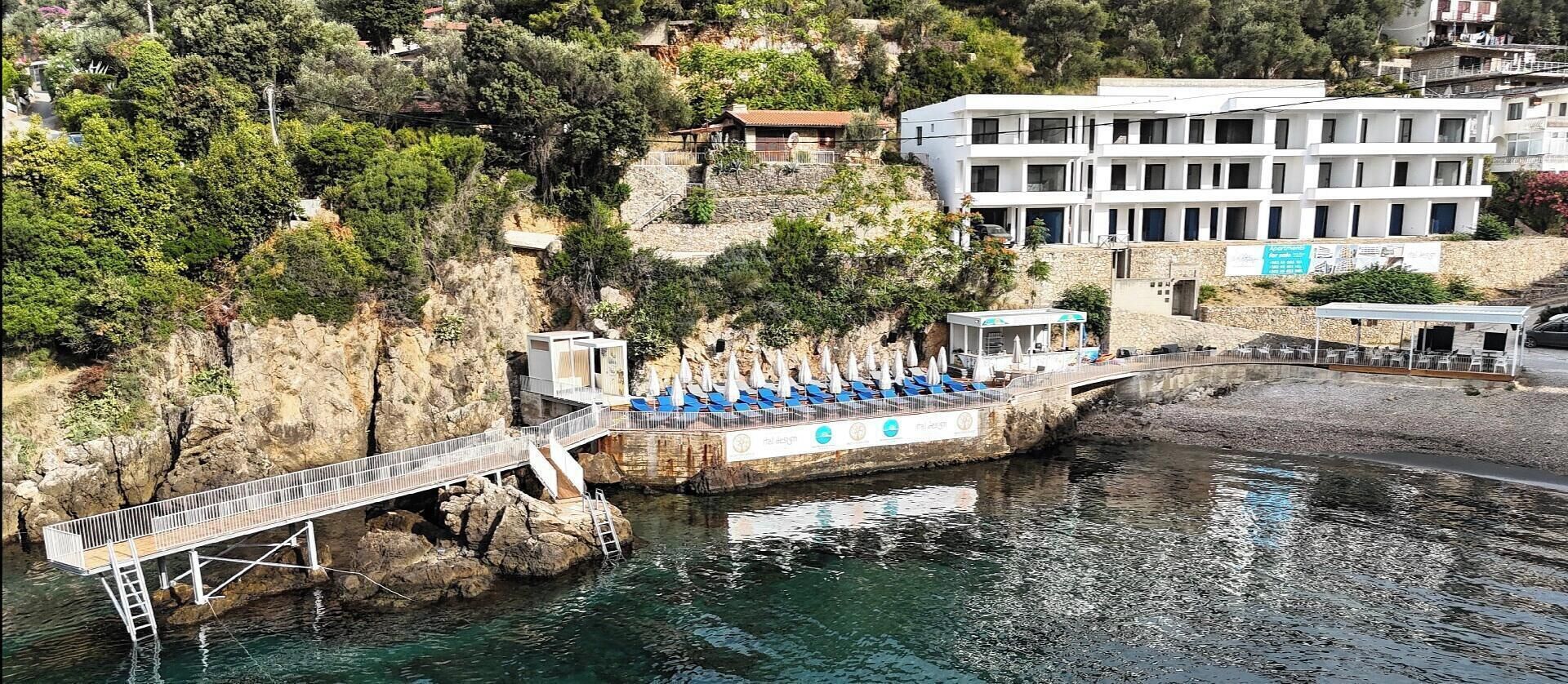 A coastal hotel with a rocky cliff, a bridge, and a pool area with umbrellas.