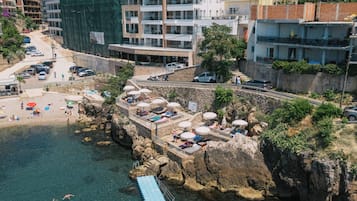 Un hotel costero con varios pisos, una zona de playa con sombrillas y un litoral rocoso.