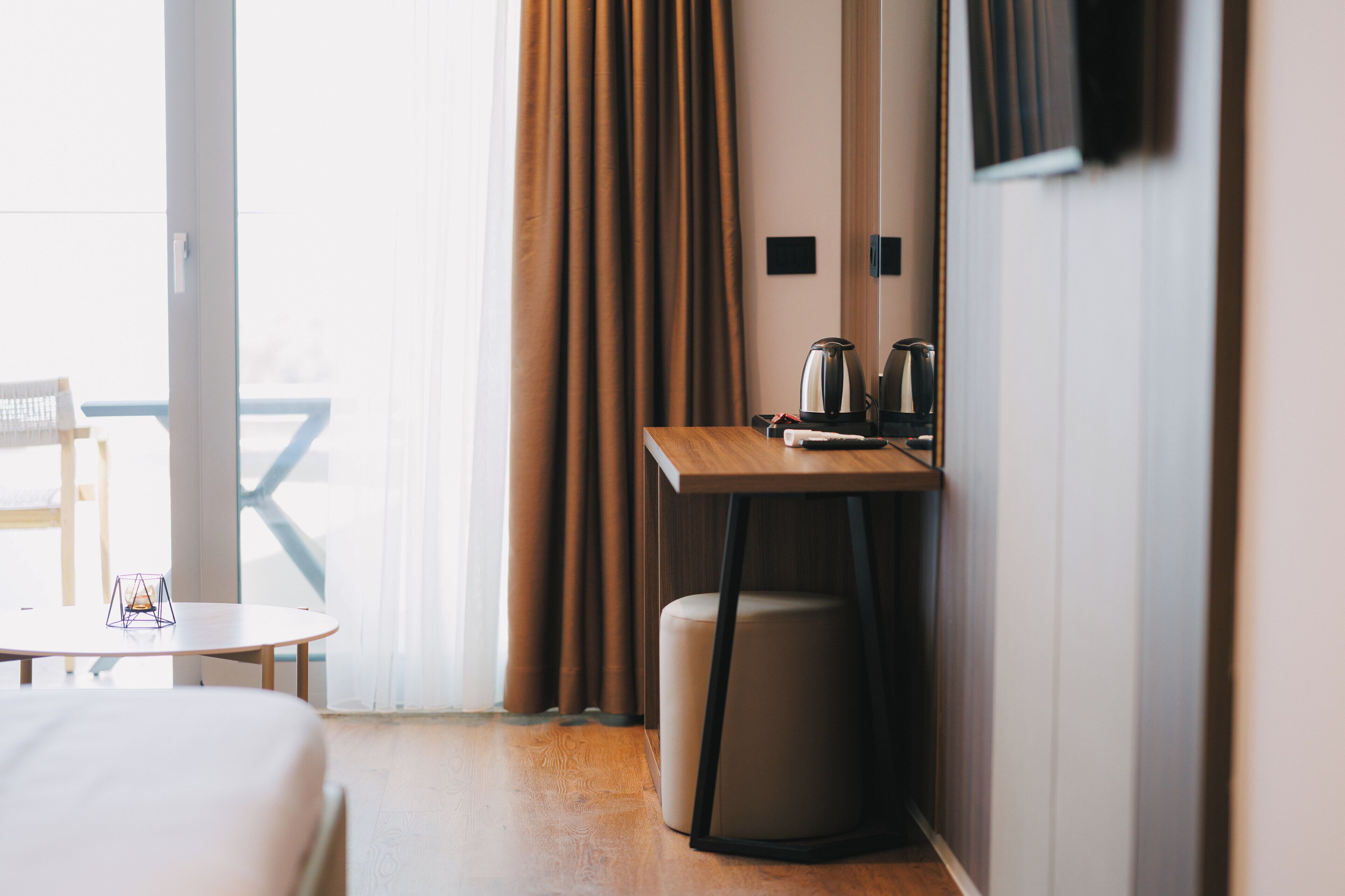 A hotel room with a bed, a small table, a chair, a desk with a kettle, and curtains.