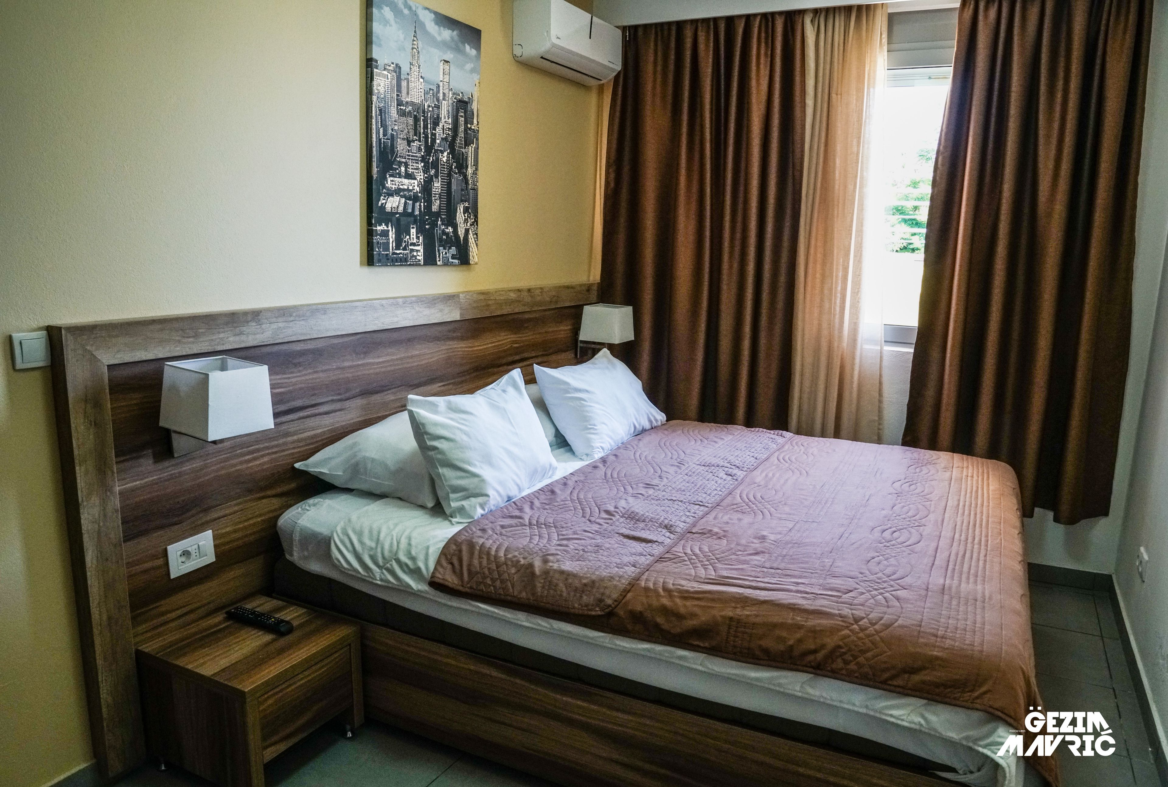 A hotel room with a wooden headboard, a cityscape painting, a brown bedspread, and a window with brown curtains.