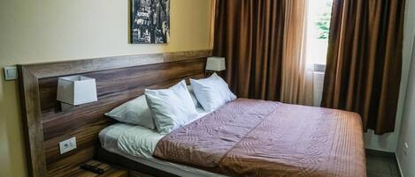 A hotel room with a wooden headboard, a cityscape painting, a brown bedspread, and a window with brown curtains.