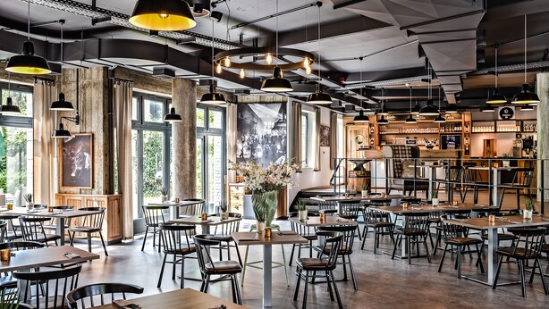 Un restaurante espacioso con techos altos, vigas a la vista y ventanas amplias. Las mesas y sillas están dispuestas para comer.
