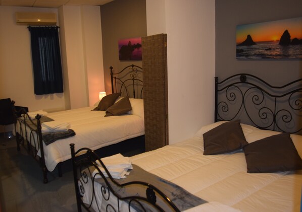 Habitación de hotel con dos camas individuales, un separador de ladrillo, un cuadro de un atardecer y una ventana con cortinas.