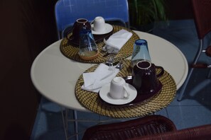 Una mesa redonda para comer con un mantel tejido, un plato blanco, una taza blanca, una tetera de color oscuro y una botella de vidrio azul.