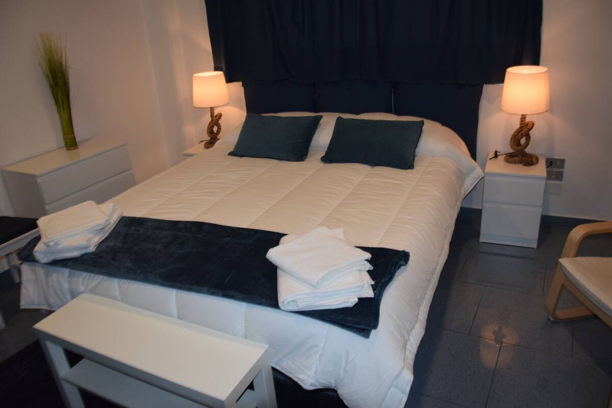 Un letto rifatto con lenzuola bianche e blu scuro, due abat-jour, un comodino e una sedia.