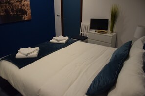 Una cama bien hecha con ropa de cama blanca y azul, una mesita de noche con televisor de pantalla plana y un florero con plantas verdes, y una pared azul con un cuadro enmarcado.
