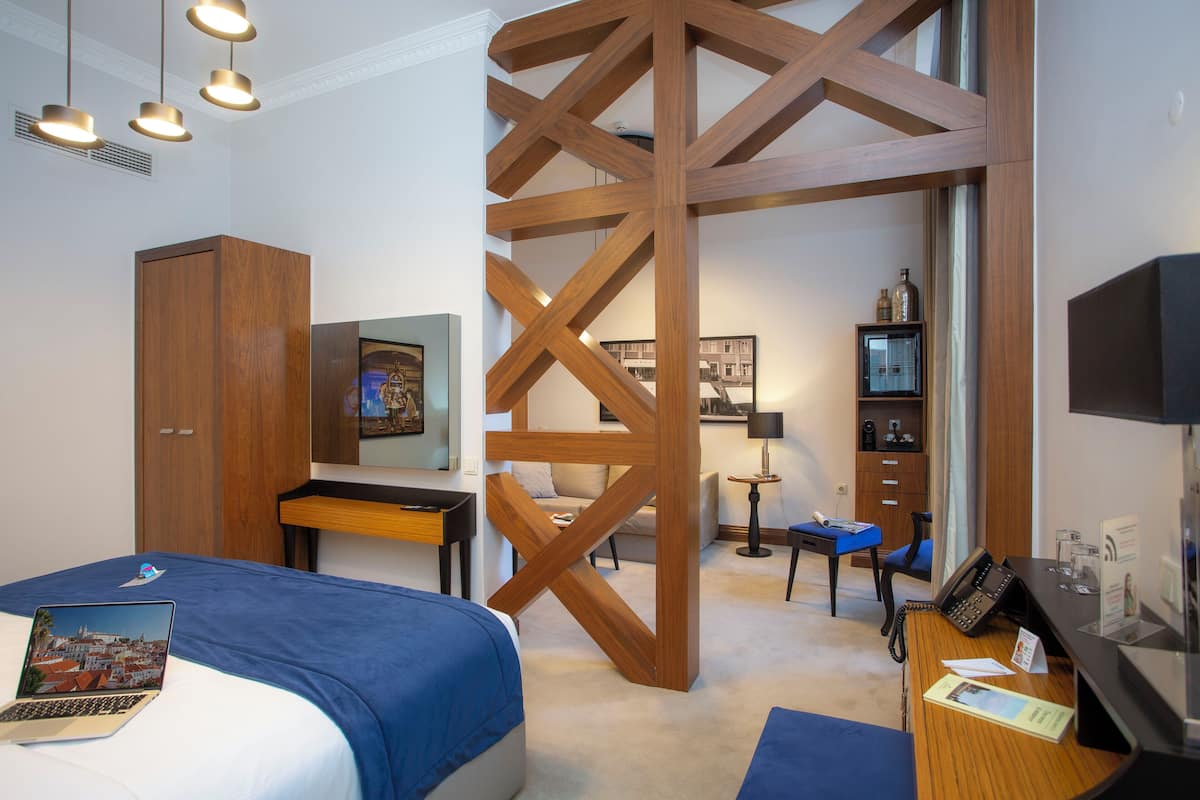 A modern hotel room with a wooden accent wall, a bed with a blue bedspread, a desk with a laptop, a chair, and a small table with a lamp.