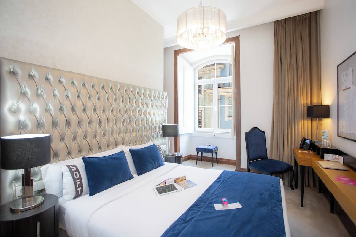 A hotel room with a tufted headboard, a desk with a chair, a lamp, and a bed with blue bedding and a magazine.