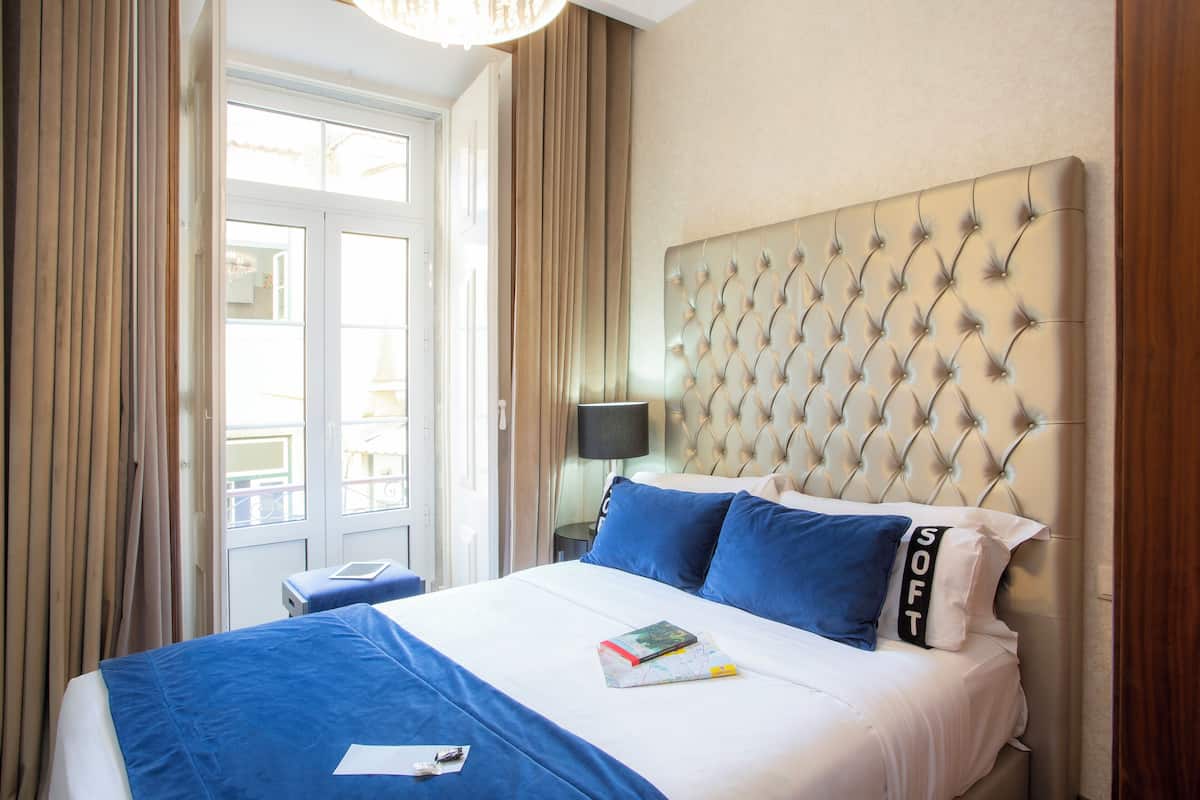 A hotel room with a large bed, blue bedding, a tufted headboard, and a view through a window with curtains.