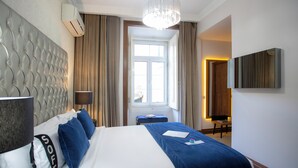 Ein Hotelzimmer mit einem großen Bett, einem Kopfteil, einem Nachttisch, einer Lampe, einem Fenster mit Vorhängen, einem an der Wand befestigten Fernseher und einer Chandelüster.