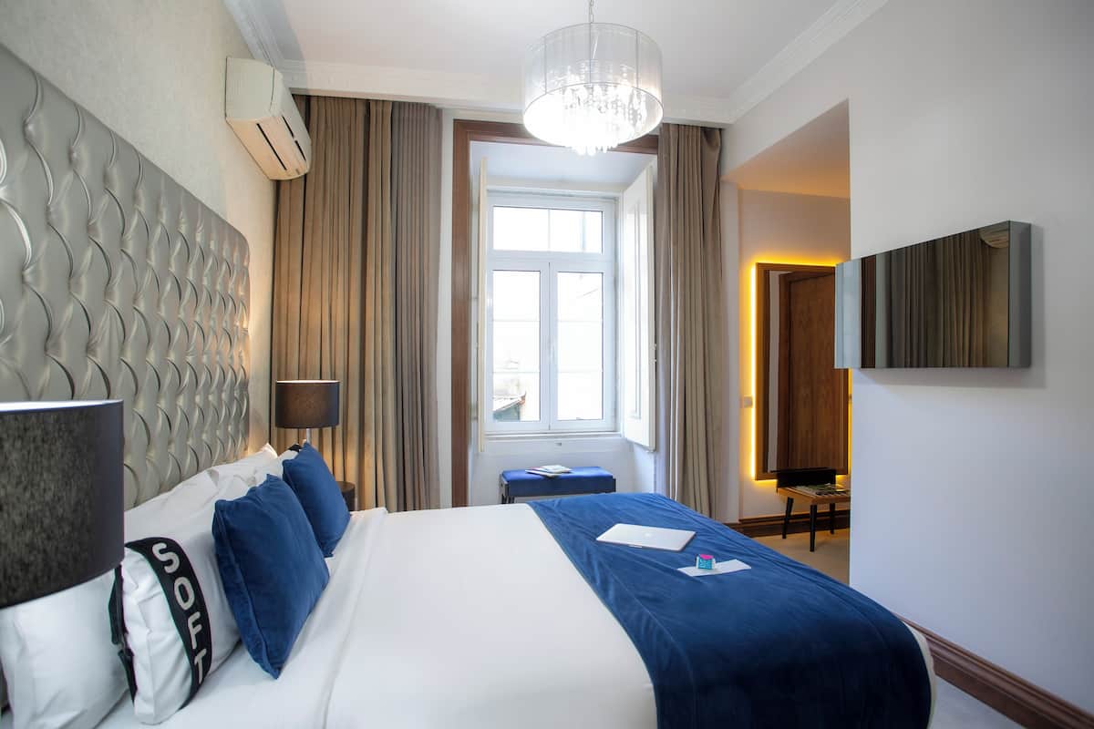 A hotel room with a large bed, a headboard, a bedside table, a lamp, a window with curtains, a wall-mounted TV, and a chandelier.