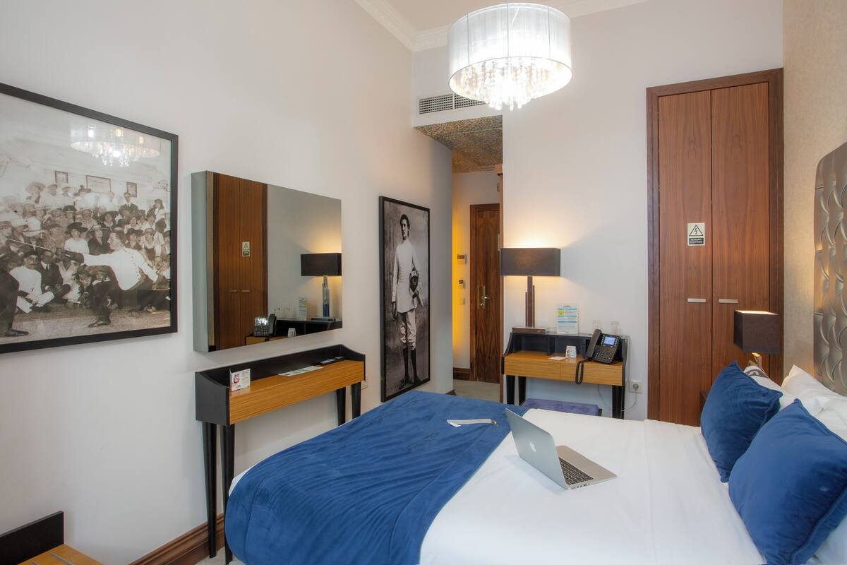A hotel room with a bed, a desk with a laptop, a mirror, and a framed photograph on the wall.