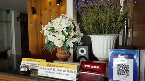 Un mostrador de recepción con un florero de flores blancas, una planta en maceta con flores moradas, una cámara, un buzón rojo y un cartel con los horarios de registro de entrada y salida.