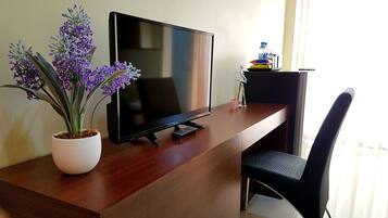 A modern hotel room with a wooden desk, a flat-screen TV, a potted plant, a chair, and a small table with a water bottle and a figurine.